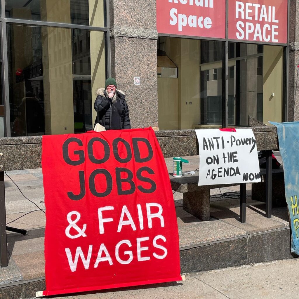Justice for Workers Nova Scotia Responds to Recommended $0.20 Minimum-Wage Increase-Photo-A speaker at a rally stands behind a large red sign that says "Good Jobs & Fair Wages" next to a smaller white sign that says "Anti-Poverty On the Agenda Now"